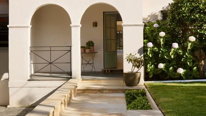 Image of the front of a white house that has two archways leading to a front stoop with a sage green door that is halfway open. There is a stone walkway and a grassy front yard