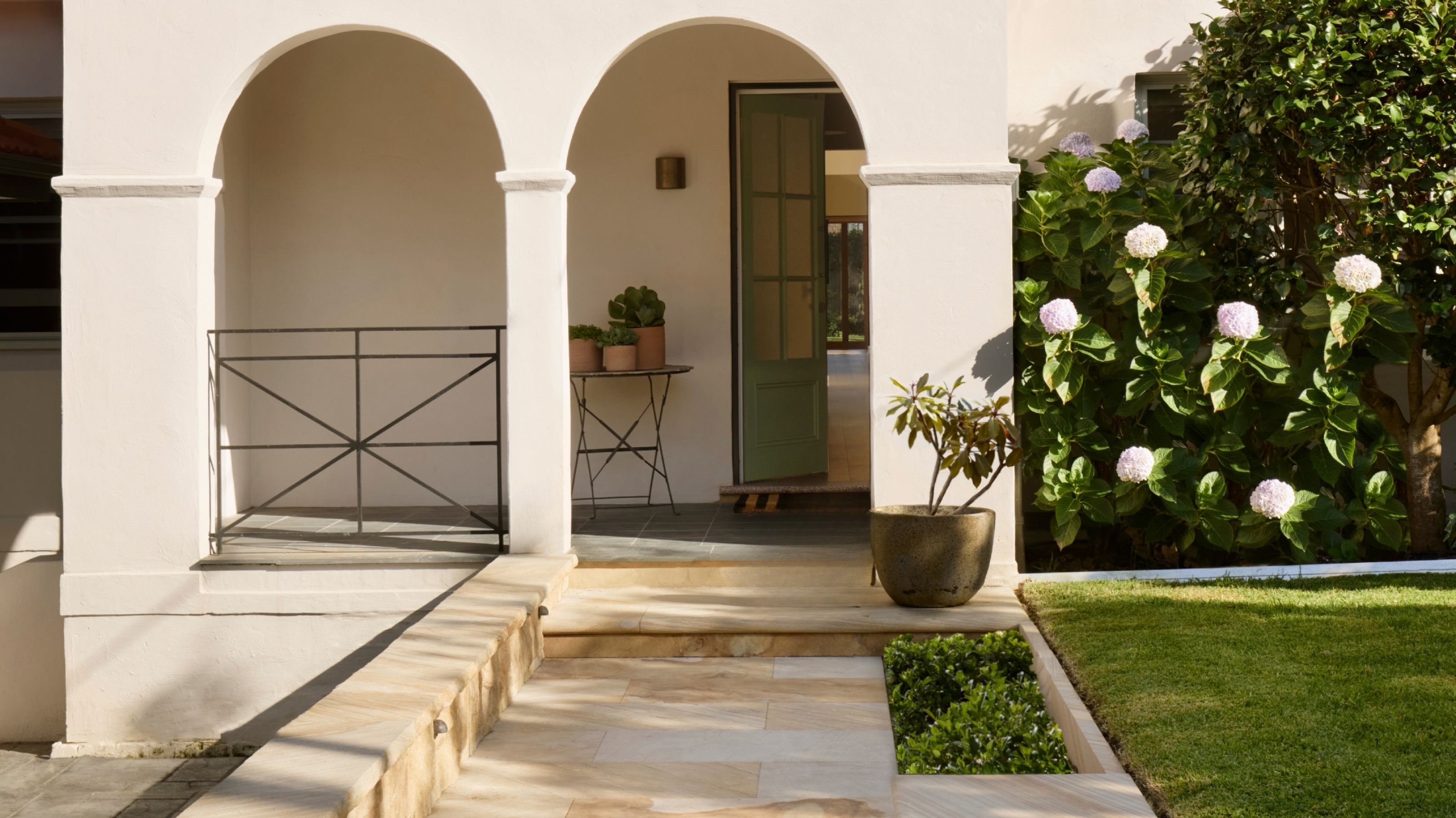 Image of the front of a white house that has two archways leading to a front stoop with a sage green door that is halfway open. There is a stone walkway and a grassy front yard