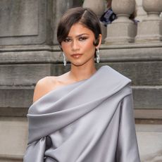 Zendaya poses for a photo wearing grey pointed-toe heels with a grey satin minidress and drop earrings.
