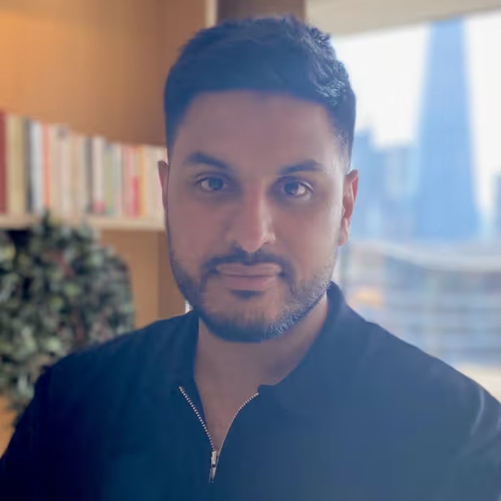 A headshot of a man with short dark hair and a beard wearing a black zip-neck top, with a bookshelf and city skyline window visible behind him.
