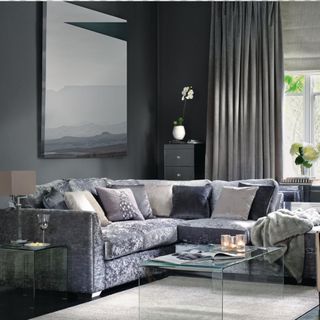 Modern grey living room with L shaped grey crushed velvet sofa and minimal glass coffee table on cream rug.