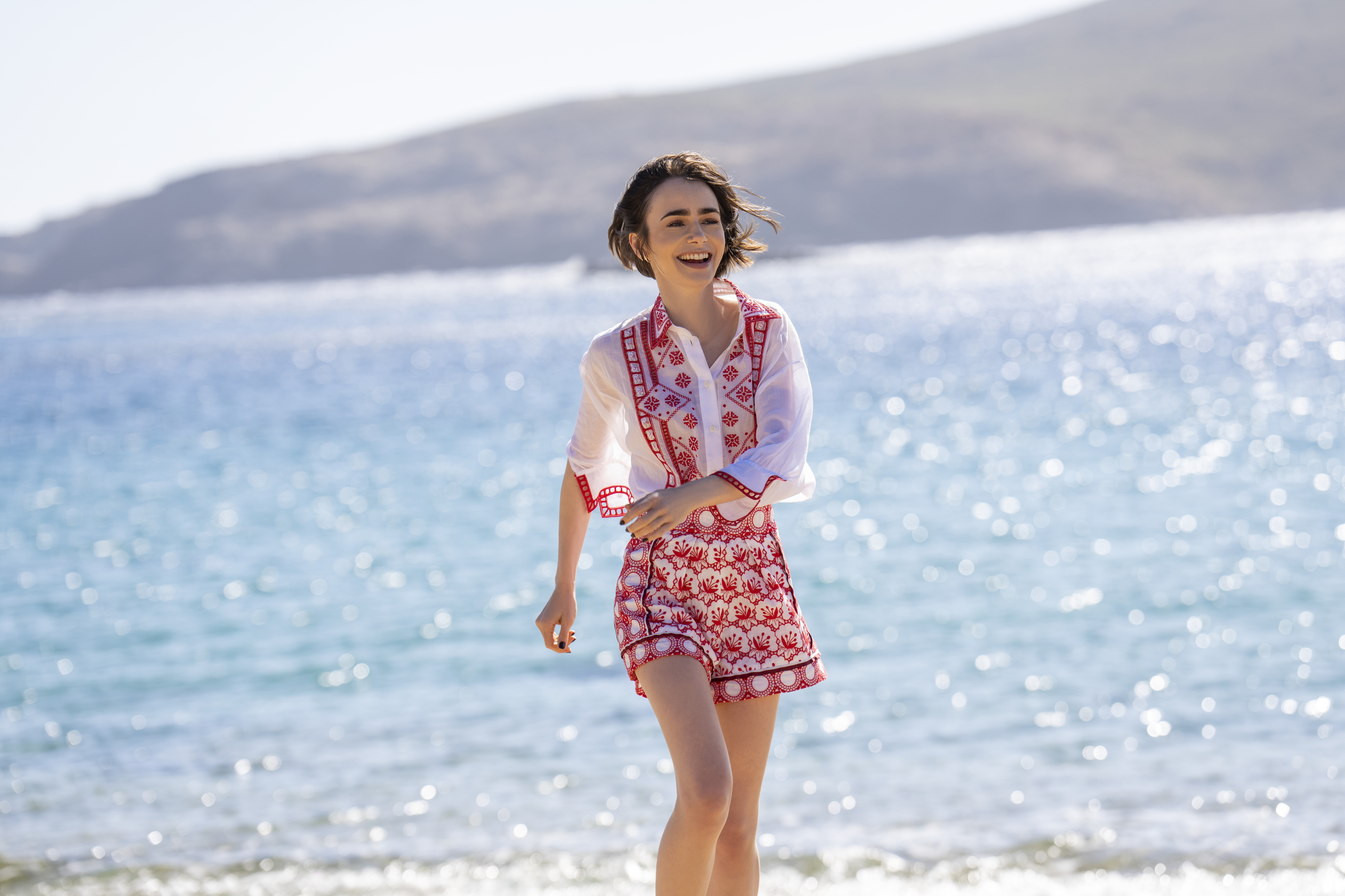 Lily Collins as Emily wearing a white and red outfit running on a sunny beach in greece in Emily in Paris season 6