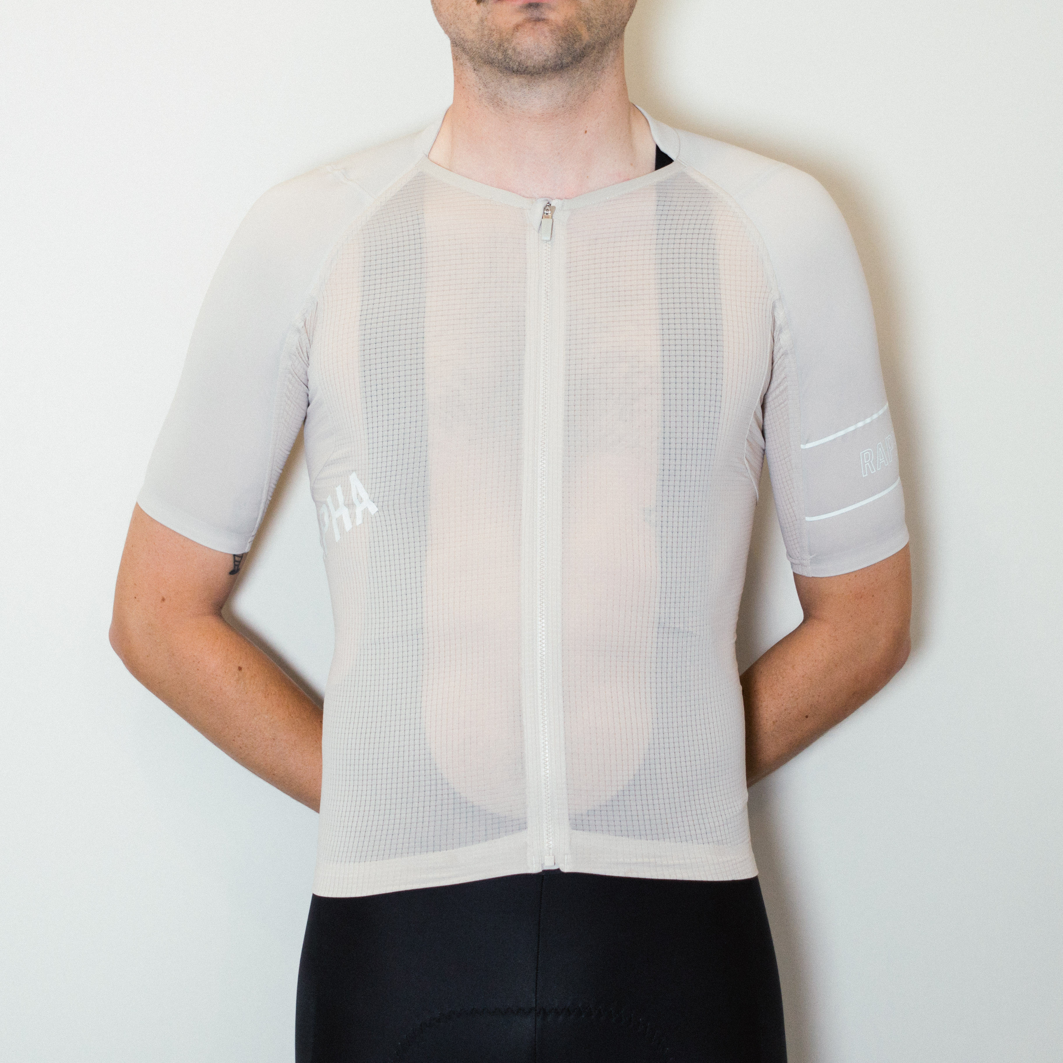 A white man in a beige cycling jersey and black cycling shorts stands against a white wall