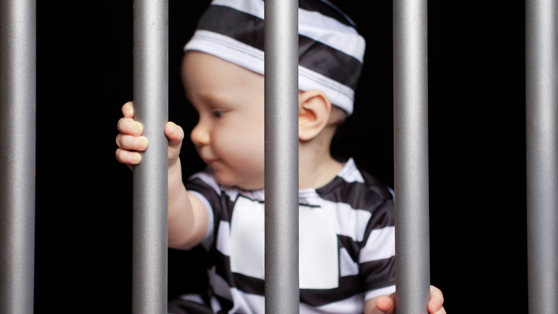 A baby dressed in prison stripes behind bars.