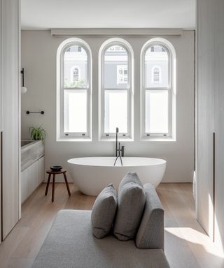 white bathroom with arched windows, freestanding bath, gray sofa and wooden floor