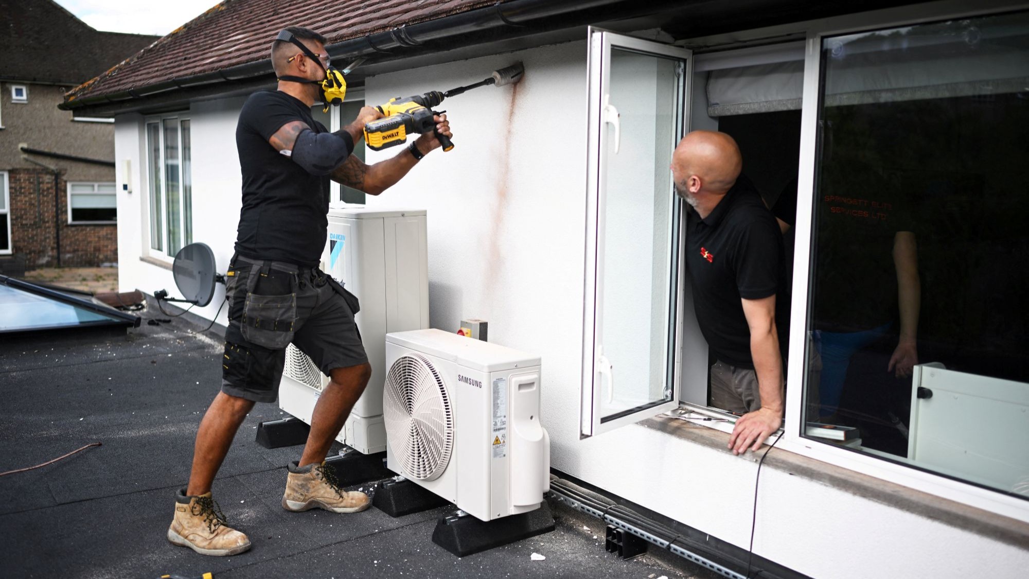 A air to air heat pump being installed on a roof with a workman drilling a hole in the wall for the air conditioning unit