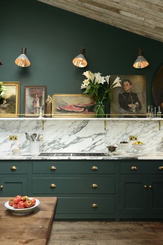 A kitchen with dark green cabinets, a marble splashback, a shelf with vintage paintings with wall sconces above it