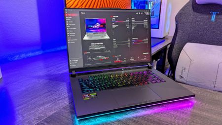 A gaming laptop with a colorful backlit keyboard displays system information on the screen, set against a modern workspace with ambient lighting.
