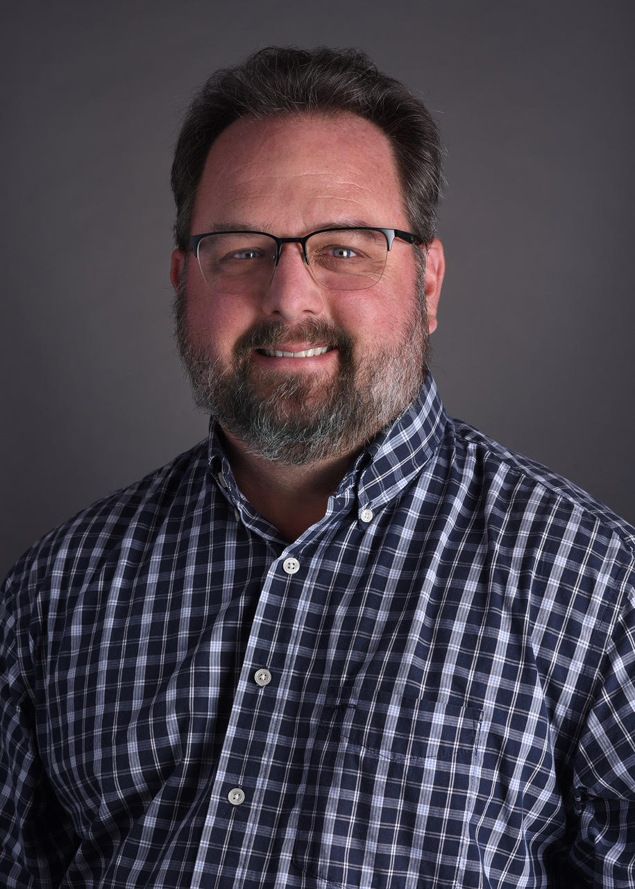 Headshot of male in blue check shirt, glasses and a beard