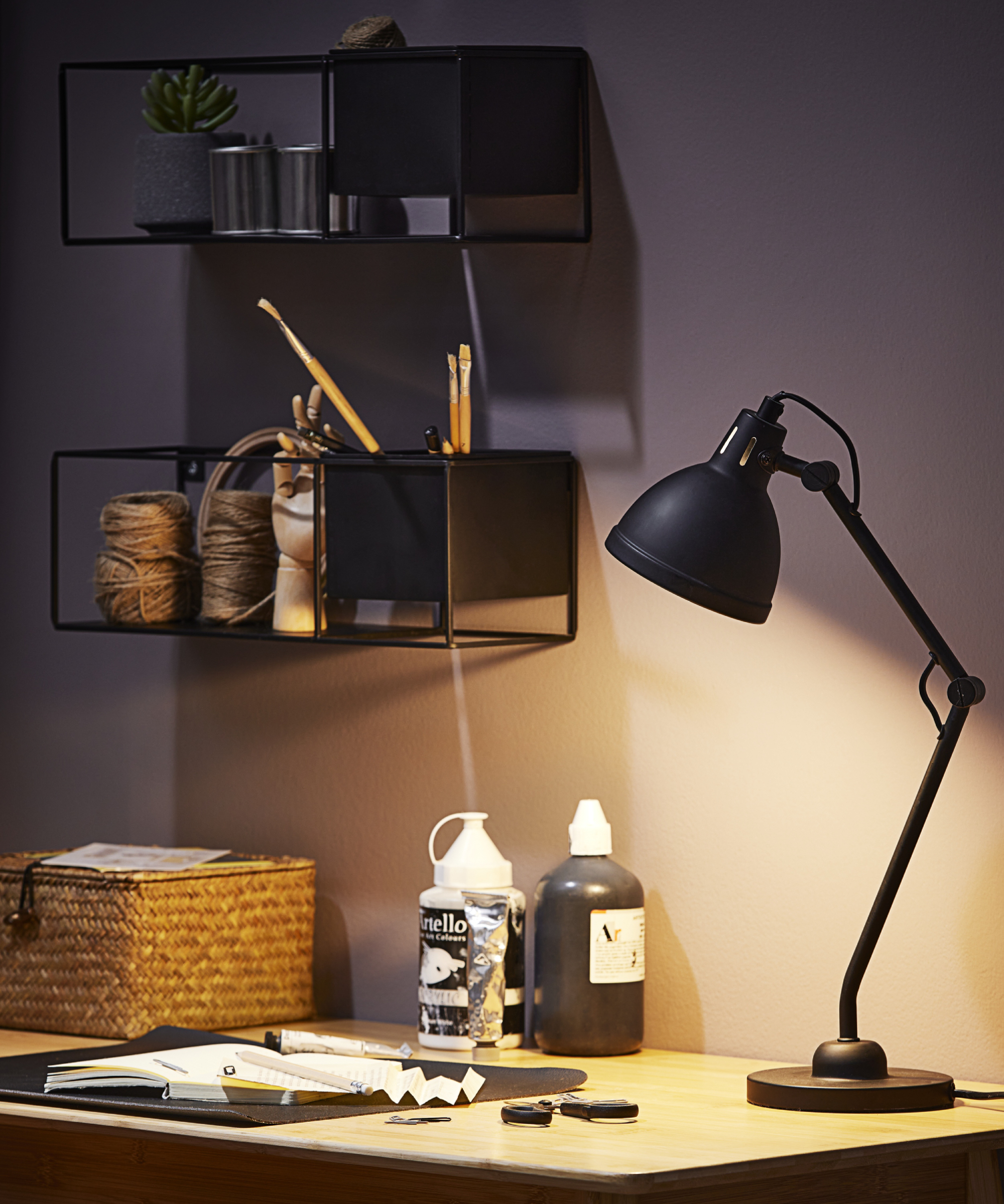 black desk lamp on wooden desk with black metal shelving above desk, art supplies on desk