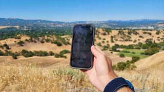 The author holding an iPhone 14 Pro above a hilly region in direct sunlight. The AllTrails app tracking his location is barely visible.