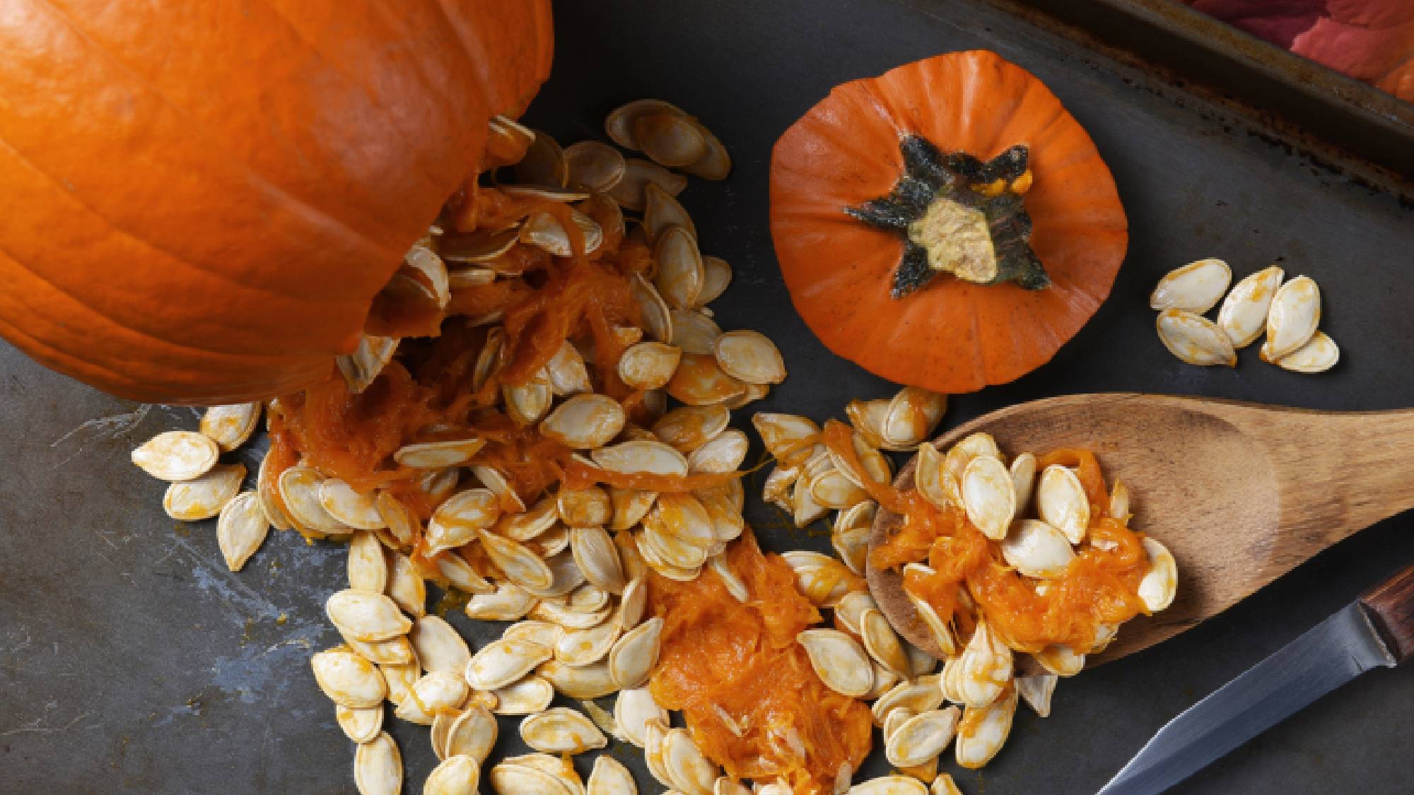 Raw pumpkin seeds and stringy orange pulp spilled on a metal baking sheet next to a wooden spoon and paring knife.