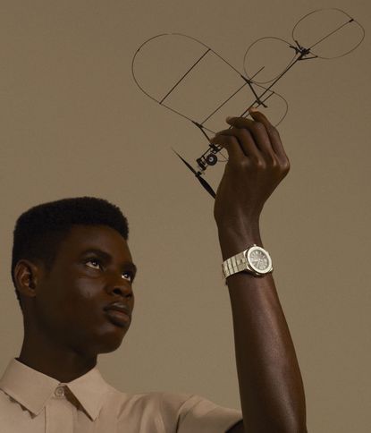 An image of a black male looking up to arm with a wrist-watch on. The watch is silver in colour with a circular navy face and Roman numerals at 12, 3, 6 and 9. Line marks cover the other numbers. 