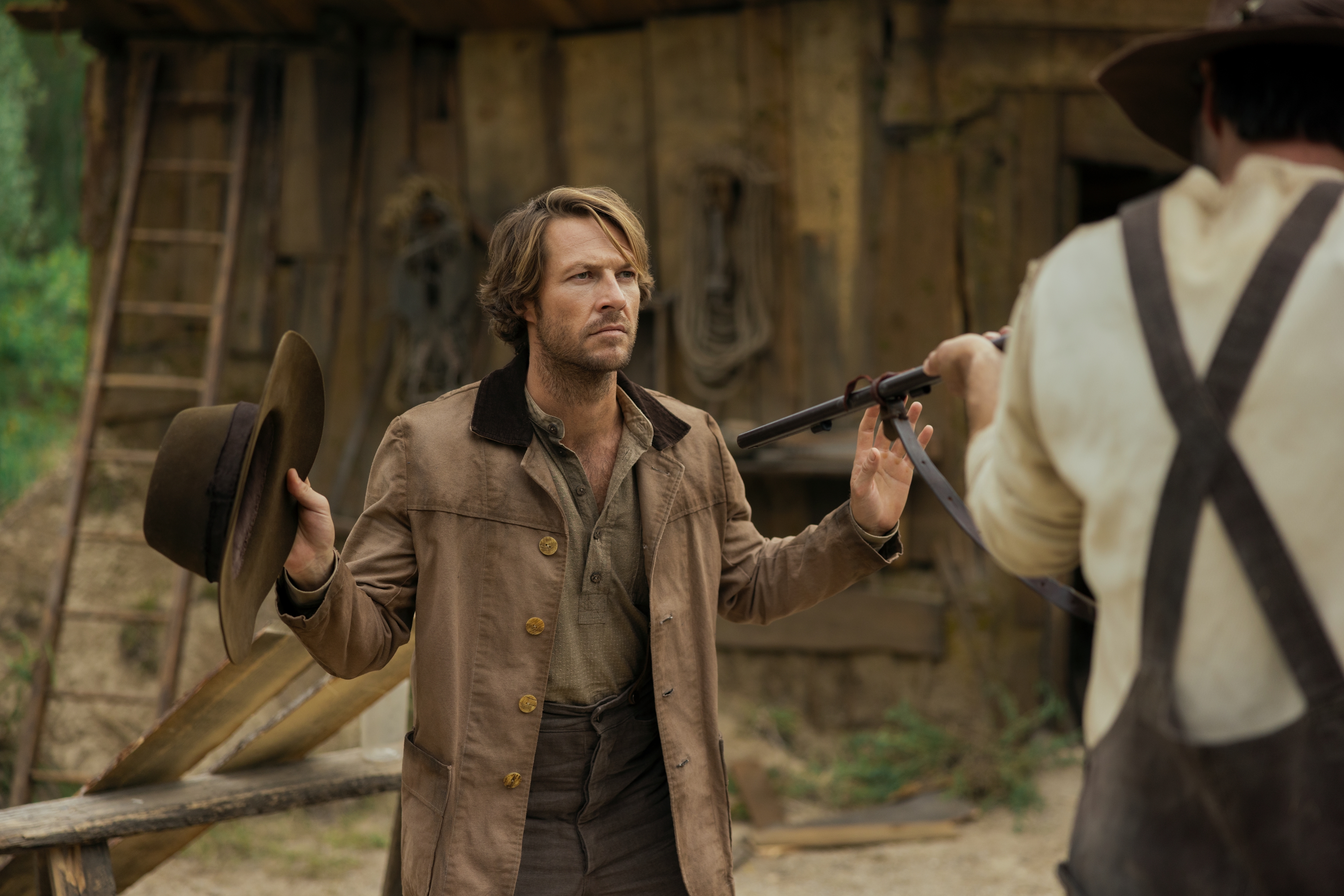 Actor Luke Bracey as Charles Ingalls, posing with his hands up while holding a hat, as an unseen man points a rifle at him, in Netflix's 'Little House on the Prairie.'