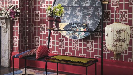 Wall decorated with bright pink chinese style wallpaper and pink skirting board, dark wooden flooring, a colourful rug and black metal bench