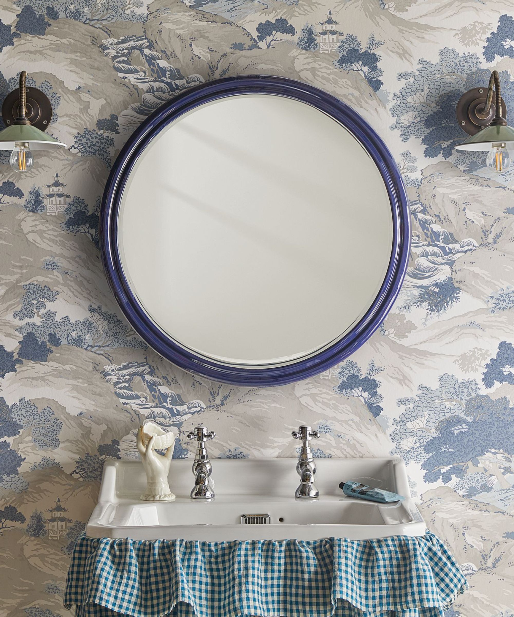 A bathroom sink with a small blue gingham curtain underneath, a circular mirror above with a blue frame and two scone lights either side.