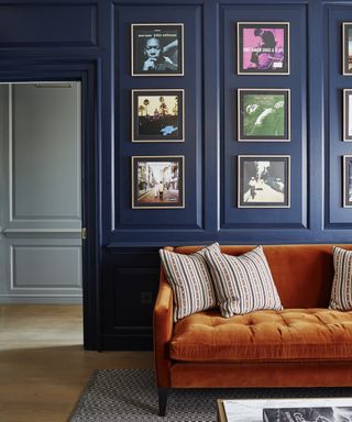 A blue living room with navy blue panelled walls, a burnt orange velvet sofa and a gallery of wall-mounted vinyl album covers.