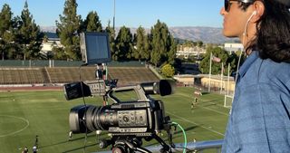 A camera man looks on a high school football field using a JVC camera on a bright, sunny day.