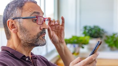 An older man wearing glasses looks closely at his smartphone.