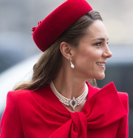 Kate Middleton wearing a red hat and coat with pearl necklace turning to the right and smiling