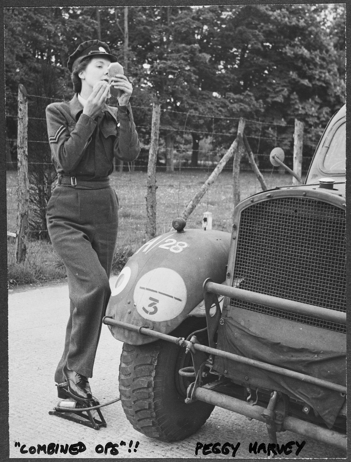 'Combined Ops!' - WAAF Corporal Peggy Harvey, in her uniform, doing her makeup whilst pumping up the tyre of a military vehicle at RAF Iver Heath, Pinewood Studios