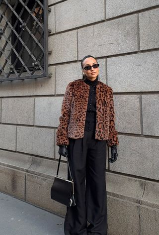 A woman wearing black sunglasses, gold earrings, a leopard print fur coat, a black cardigan, black trousers, black leather gloves, and a black Frame bag.