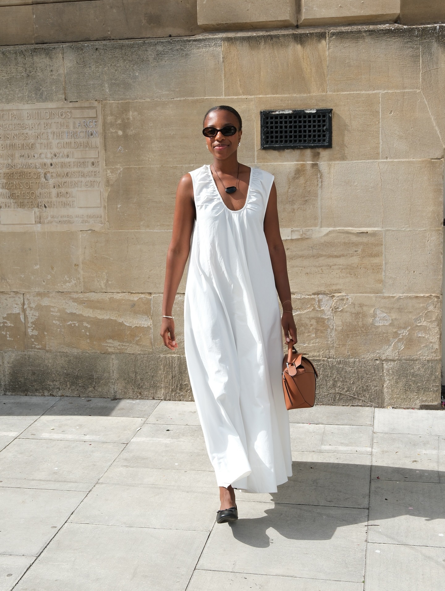British Taffy Msipa poses in black oval sunglasses, a black cord pendant necklace, ruched white sleeveless dress, tan Loewe puzzle bag, and black ballet flats