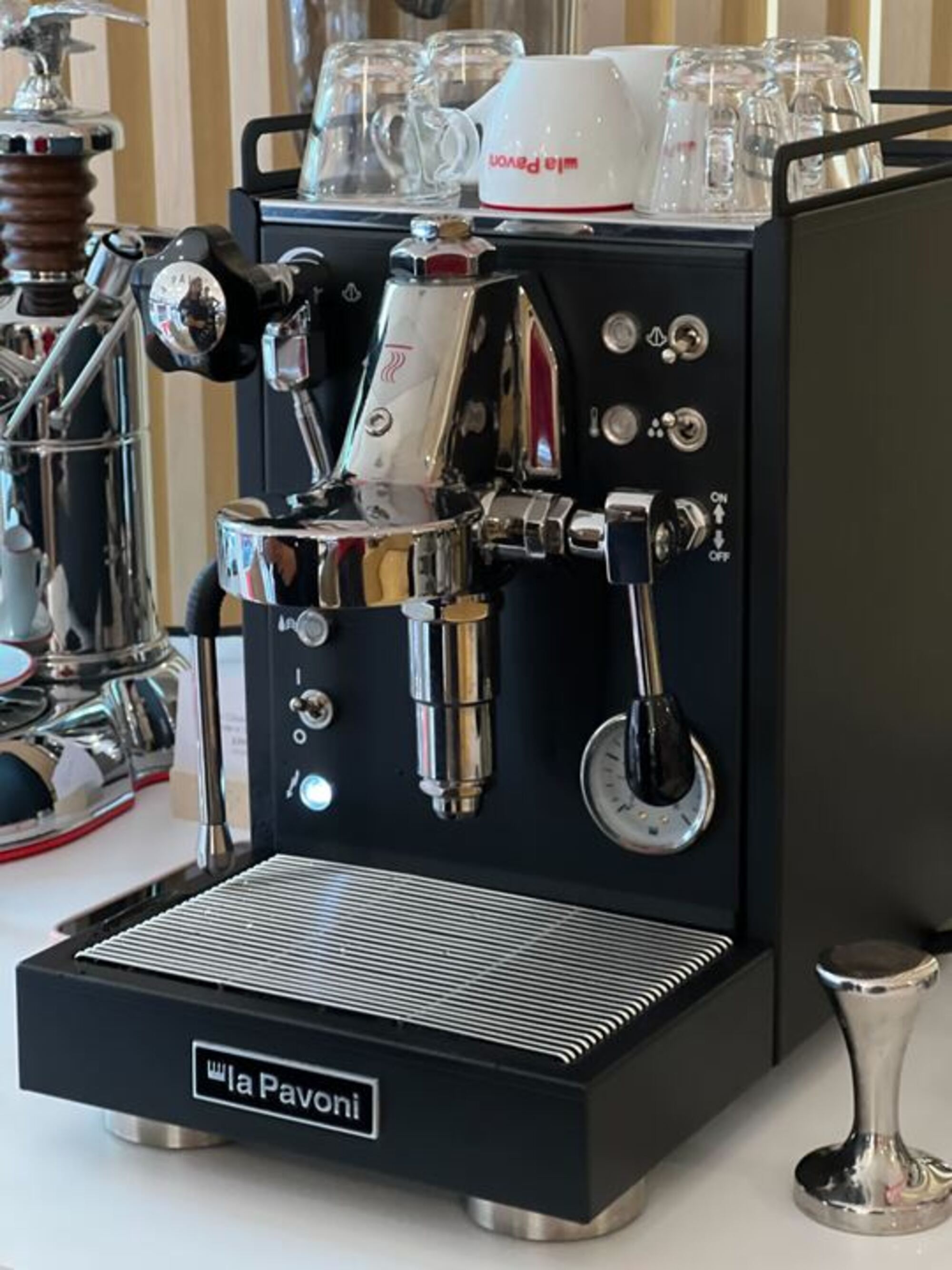 Image of a black La Pavoni Mini Cellini Semi-Professional Domestic Coffee Machine on a white countertop with coffee mugs on the top of the machine