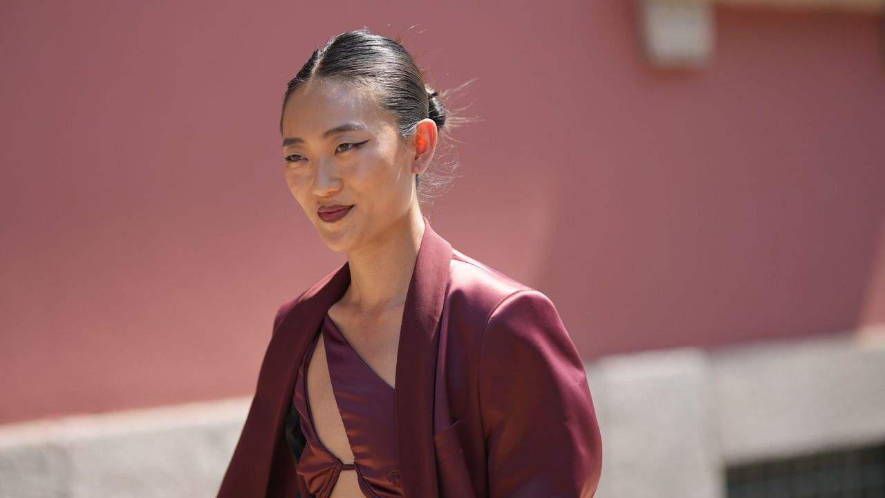 best brown eyeliners - woman smiling wearing a burgundy top and blazer with a chignon and a sharp brown eyeliner flick and burgundy lip - gettyimages 1499115748