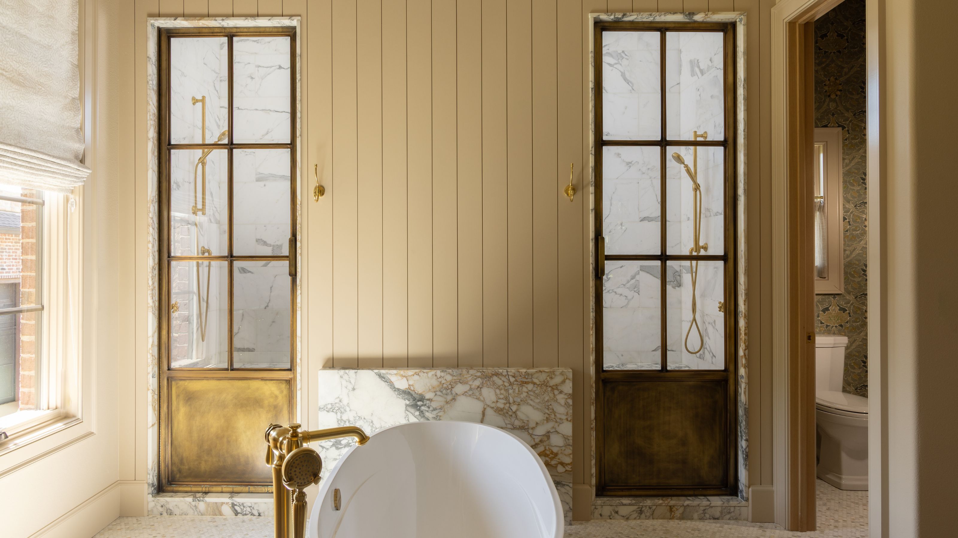 a beige painted and paneled primary bath with freestanding tub and two brass crittal shower screen doors for dual entry