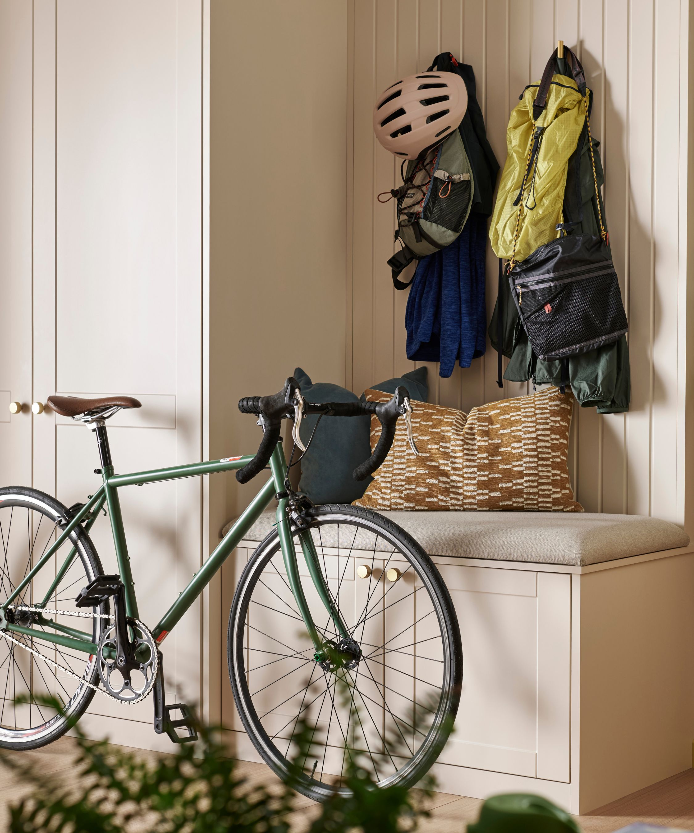 A cream hallway with tongue-and-groove panelling, a built-in storage bench with brass knobs, wall hooks holding a cycling helmet and backpacks, and a green road bike.