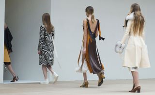 Three models on a catwalk, one wearing a zebra print dress, one in a brown, caramel and white dress and one in a cream coat with fur cuffs and collar