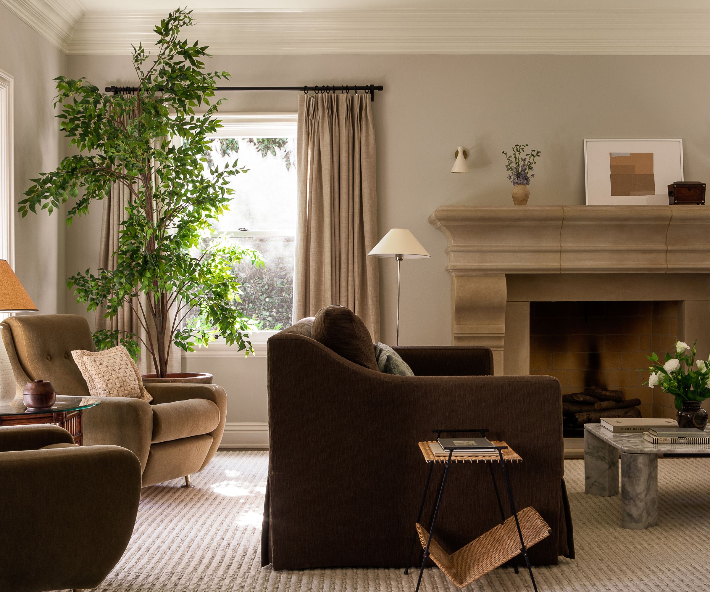 A transitional living room with pale greige walls, a large stone fireplace, beige curtains, a large chocolate brown sofa, neutral striped carpet, a large plant, and neutral armchairs.