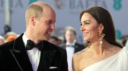 Prince William wears a tuxedo while his wife Kate Middleton wears a one-shoulder white gown with long dangly gold earrings and long black gloves