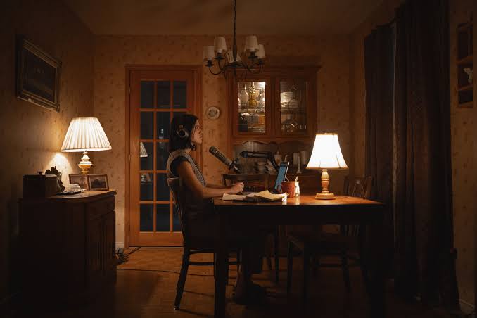 a woman sits at a desk in front of a microphone reading from a script in an old dark house in the movie undertone