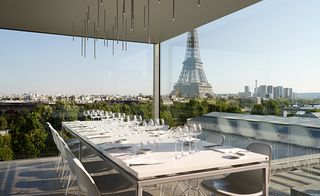 Daytime, outside view from the Nomiya Restaurant, Palais Tokyo, Paris, long dining table with row of empty drinking glasses and cutlery, chairs, Eifel tower and surrounding area in the backdrop, pale blue-sky