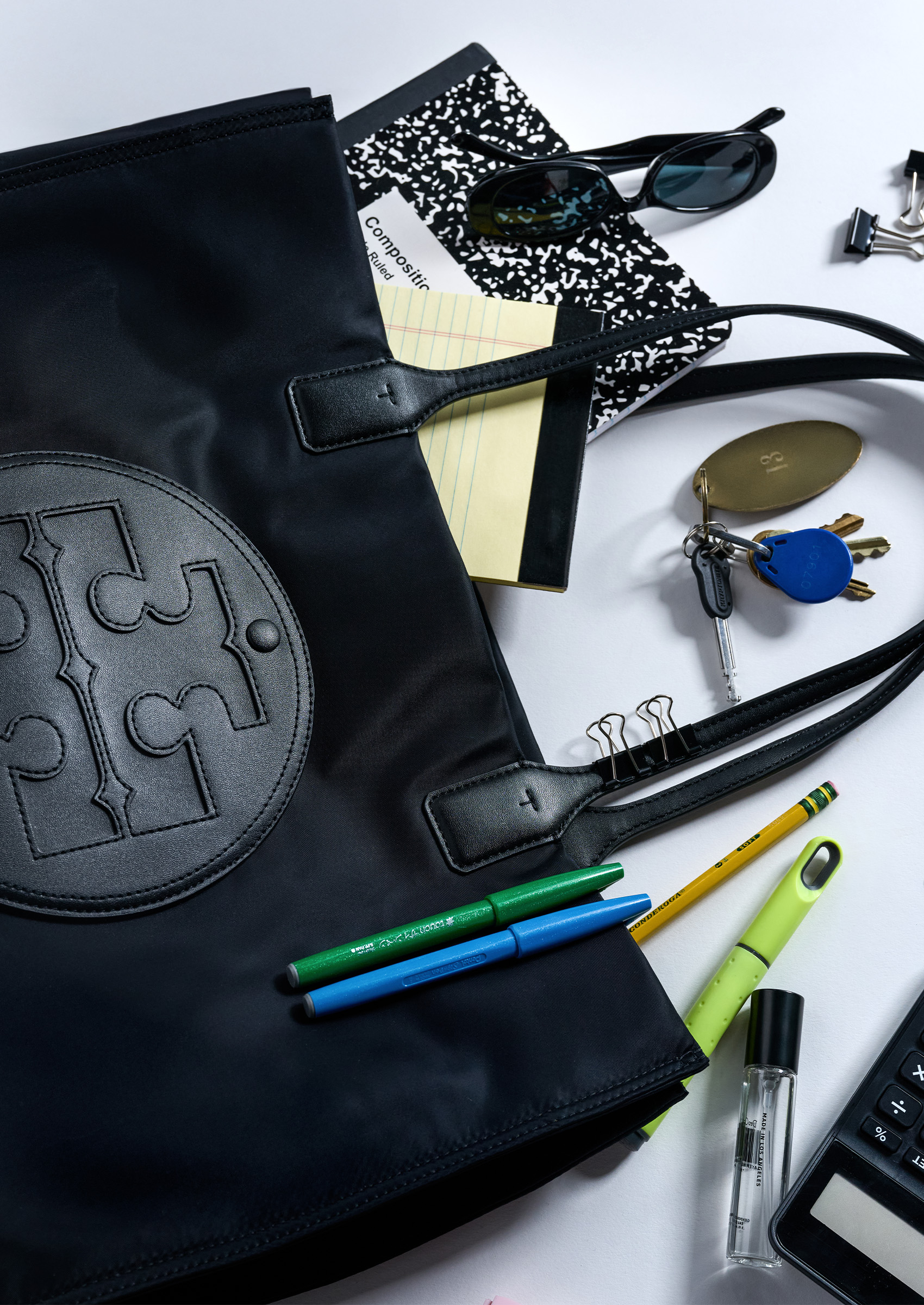 A black Tory Burch bag on a white table with notebooks, keys, pens, and pencils surrounding it