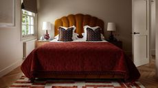 Image of a bed with a burnt orange headboard and read bedding in a small, beige bedroom. There are side tables on either side of the bed with lamps on them and a window on the left wall.