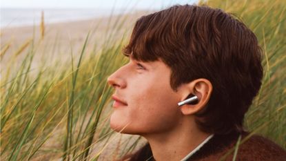 A young man wearing Bang & Olufsen Beo Grace earbuds in long grass on a beach on a cloudy day
