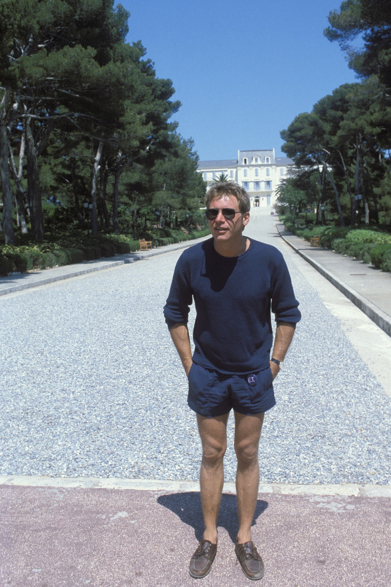 Harrison Ford in 1982 standing outside the Hotel du Cap-Eden-Roc wearing a blue sweater and blue shorts as well as sunglasses. His hands are in his pockets.
