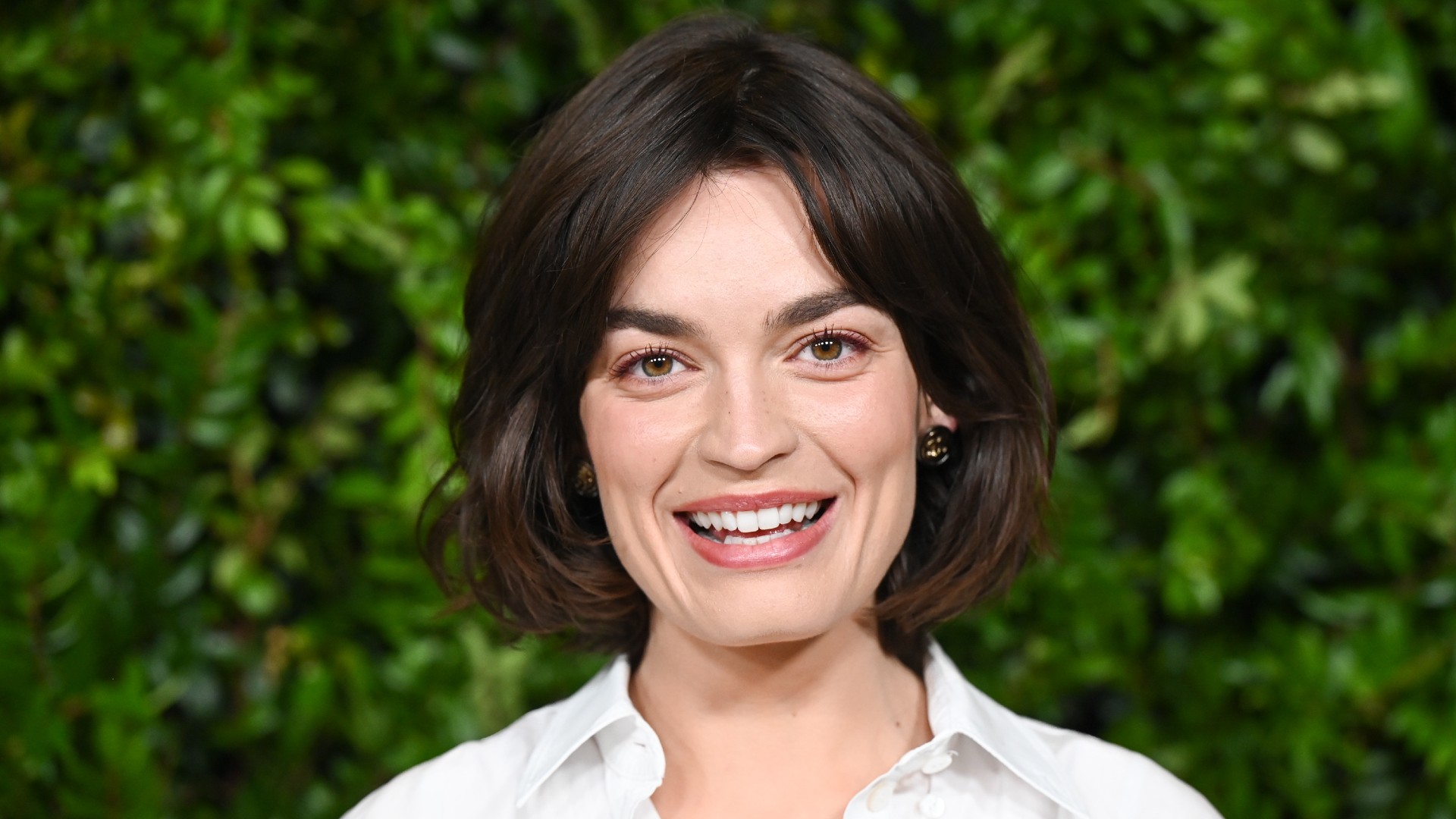a white woman with short, dark hair posing in front of a grass background