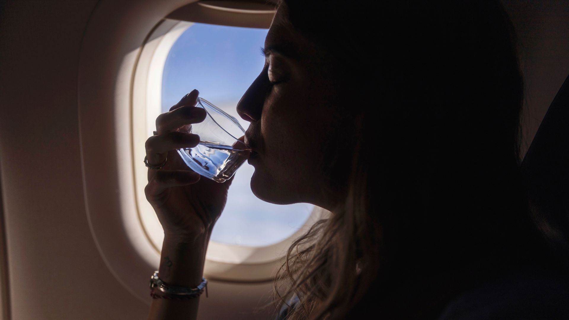 A woman with long hair takes a small sip from a small glass of water while sitting on a window seat of a plane.