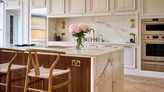 A marble kitchen island with a vase of roses on top it and dark wooden paneling on it, two wooden bar stools in front of it, cream cabinets behind it, and wooden flooring