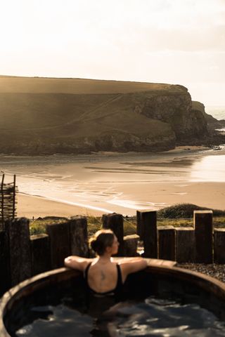 A woman sits in an outdoor bathtup while gazing at cliffs surrounded by water at sunset.