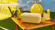 A citrus yellow Bose SoundLink Plus Portable on a wooden table with a bright yellow chair next to it and two glasses of lemonade and a big jug of lemons on the same table