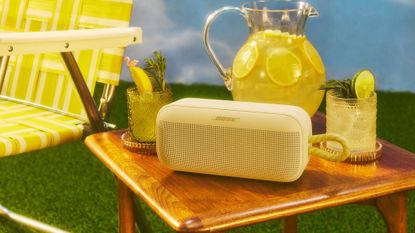A citrus yellow Bose SoundLink Plus Portable on a wooden table with a bright yellow chair next to it and two glasses of lemonade and a big jug of lemons on the same table