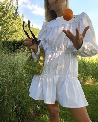 British female influencer Lucy Williams poses in the countryside wearing a short white tiered dress and small plaid bag while tossing an orange
