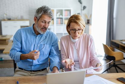An older couple sits in front of a laptop surrounded by documents, visibly pressured as they attempt to organize their finances or retirement planning.