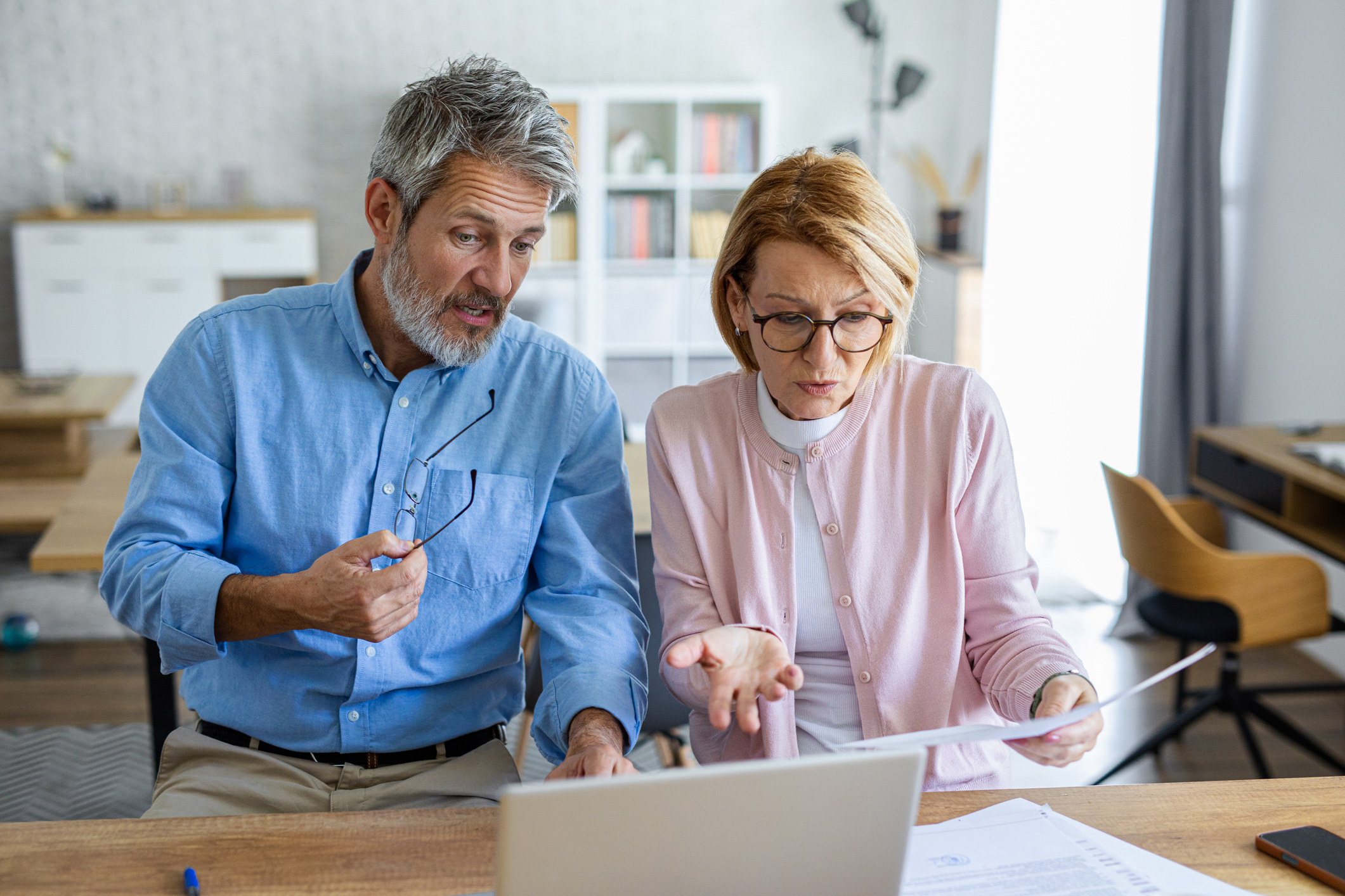 An older couple sits in front of a laptop surrounded by documents, visibly pressured as they attempt to organize their finances or retirement planning.