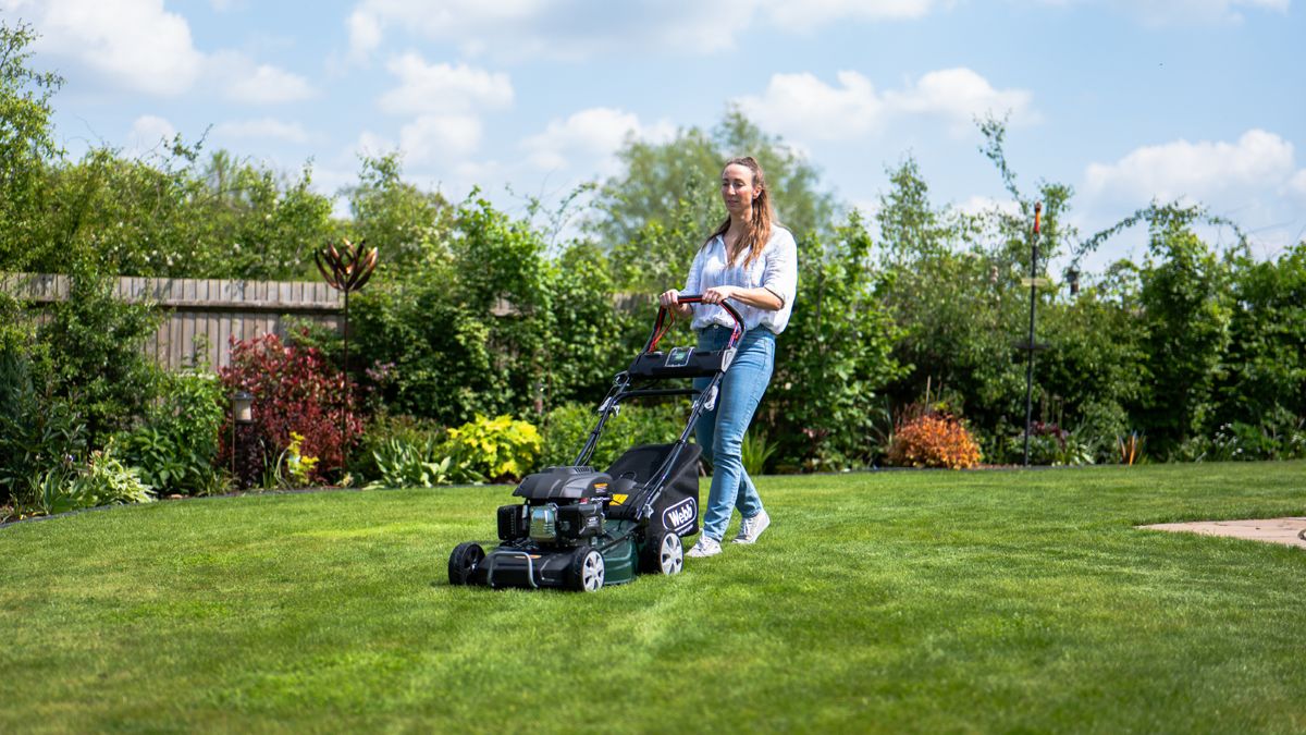 woman wearing white shirt and jeans mowing large lawn with planted borders